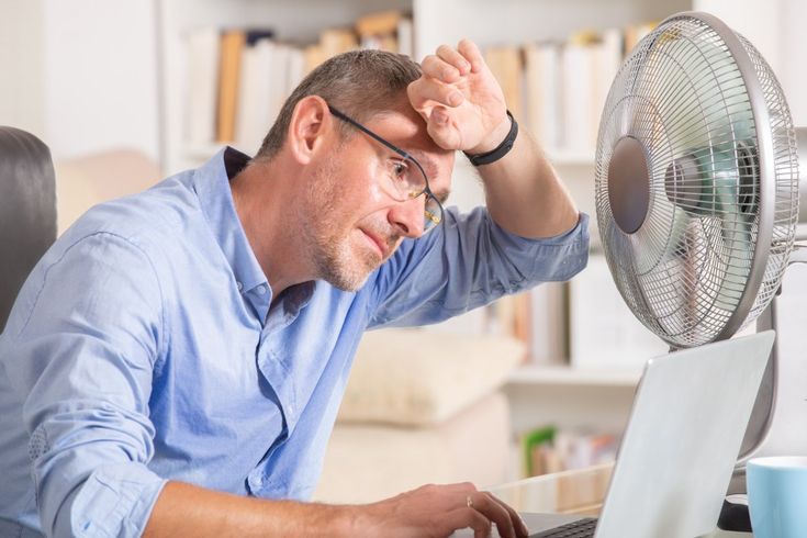 Homem se refrescando com um ventilador em frente ao notebook, sentindo o calor